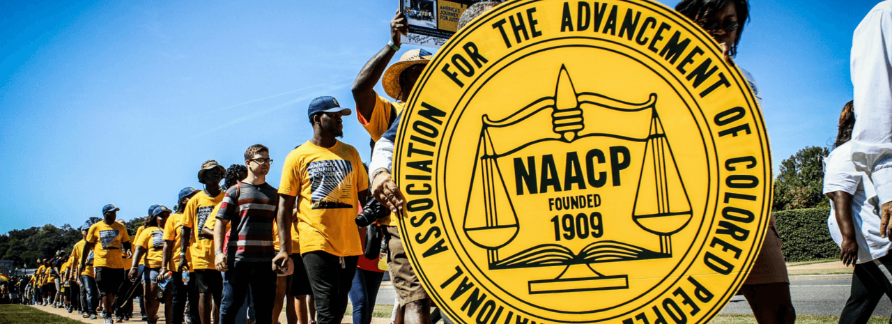 Activist holding the NAACP seal, leading a line of activists that extends out of view. They are wearing matching yellow shirts that say "America's Journey for Justice".